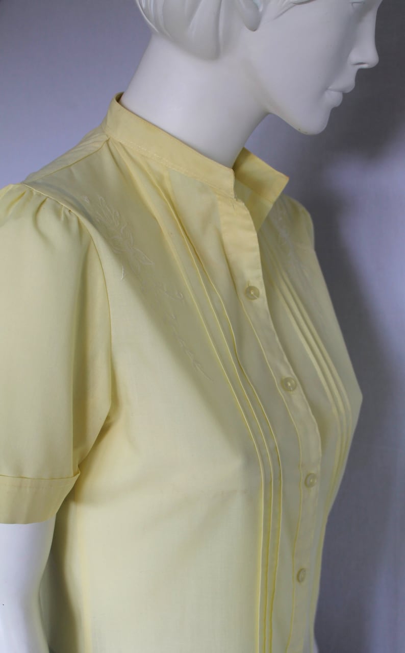 Puede incluir: Una blusa de manga corta de color amarillo p&aacute;lido con cuello mandar&iacute;n y una fila de botones en la parte delantera. La blusa tiene un frente plisado y est&aacute; hecha de una tela ligera.