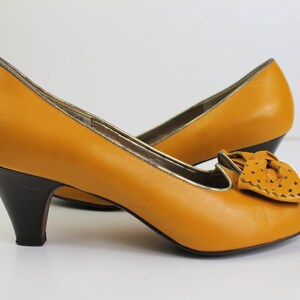 May include: Mustard yellow leather pumps with a bow detail and a patterned toe. The heels are dark brown, and the trim is gold. The shoes have a classic, vintage style.