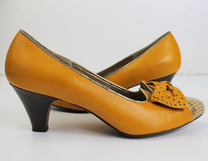 May include: Mustard yellow leather pumps with a bow detail and a patterned toe. The heels are dark brown, and the trim is gold. The shoes have a classic, vintage style.