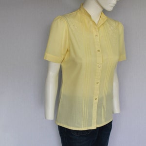 Puede incluir: Una camisa de color amarillo p&aacute;lido con botones, mangas cortas y detalles florales bordados. La camisa tiene un cuello mandar&iacute;n y un frente plisado.