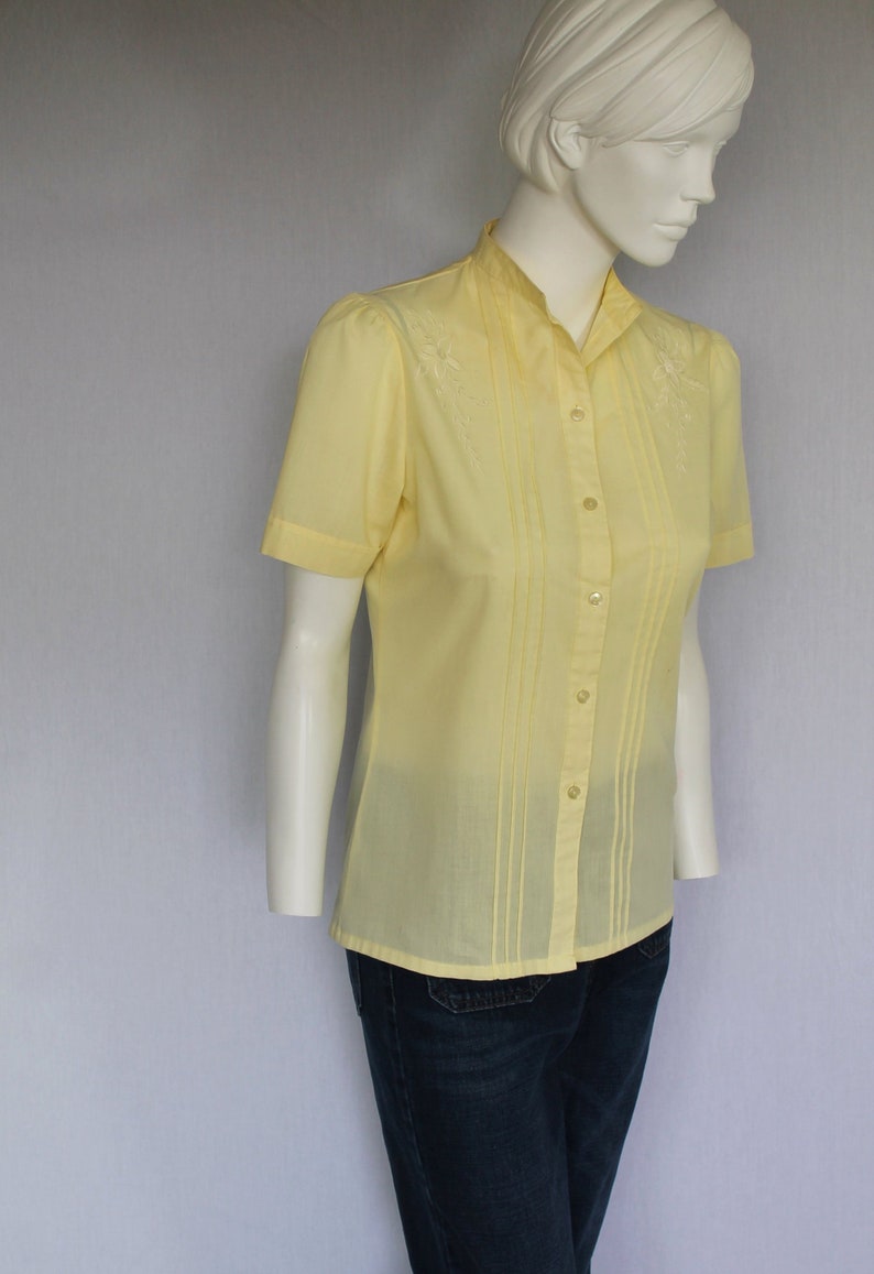 Puede incluir: Una camisa de color amarillo p&aacute;lido con botones, mangas cortas y detalles florales bordados. La camisa tiene un cuello mandar&iacute;n y un frente plisado.