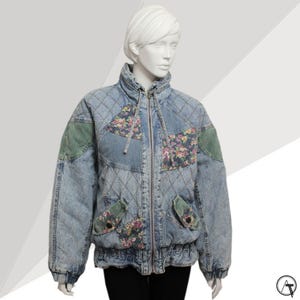 May include: A light blue denim jacket with floral and green patches. The jacket features a quilted design, a high collar, and a zipper closure. The pockets also have floral patterns and green patches. The jacket is displayed on a mannequin.