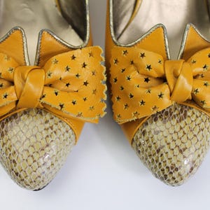 May include: A pair of mustard yellow high-heeled shoes. The shoes feature a bow with star cutouts and a snakeskin pattern on the toe. The shoes have a gold trim and a pointed toe.