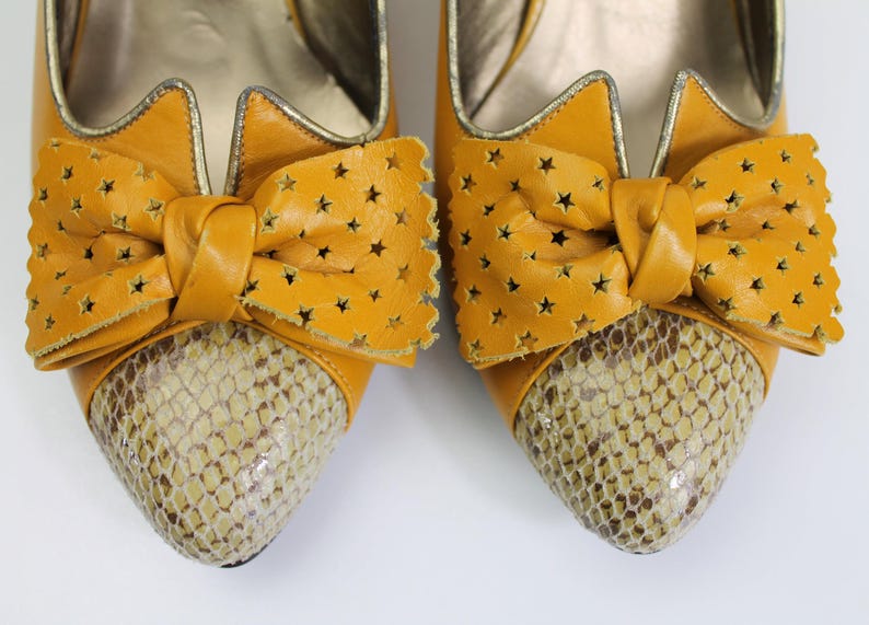 May include: A pair of mustard yellow high-heeled shoes. The shoes feature a bow with star cutouts and a snakeskin pattern on the toe. The shoes have a gold trim and a pointed toe.