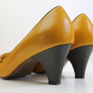 May include: A pair of mustard yellow leather pumps with dark brown heels. The shoes have a rounded toe and a small bow detail. The heels are approximately 2.5 inches high.