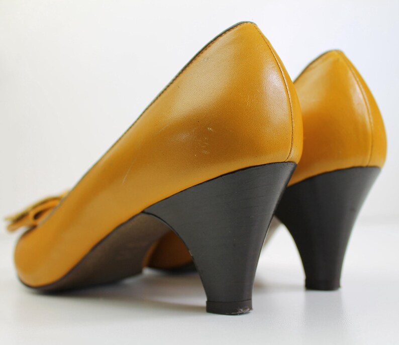 May include: A pair of mustard yellow leather pumps with dark brown heels. The shoes have a rounded toe and a small bow detail. The heels are approximately 2.5 inches high.