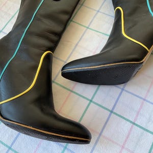 May include: Pair of black leather boots with pointed toes and green heels. The boots feature yellow and light blue trim along the seams. The soles are black with a textured pattern. A stylish and unique footwear choice.