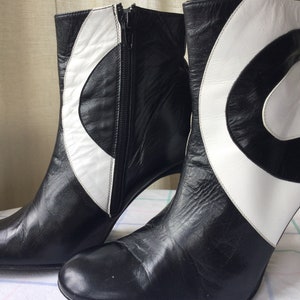 May include: A pair of black and white leather ankle boots. The boots feature a high heel and a zipper closure. A white, curved design accents the black leather. The boots are on a white and blue checkered surface.