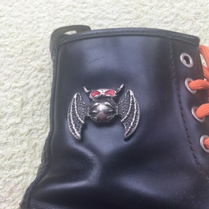 May include: Black leather boot with orange laces and a silver bat emblem. The bat has red eyes and is centered on the boot's side. The boot is laced with orange laces.