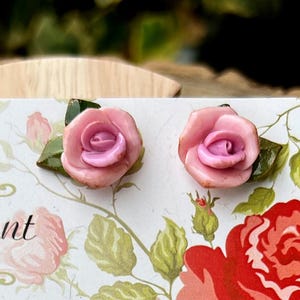Könnte beinhalten: Ohrstecker mit rosa Rosen und grünen Blättern, präsentiert auf einer Karte mit floralen Illustrationen. Handgefertigter Schmuck. Der Hintergrund ist verschwommen mit grünem Laub.