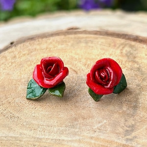 Könnte beinhalten: Ein Paar rote Rosen-Ohrstecker mit grünen Blättern. Die Ohrringe liegen auf einer Holzoberfläche mit natürlichem Rand, mit einem verschwommenen Hintergrund aus grünem Laub und lila Blumen.