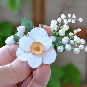 Könnte beinhalten: Eine weiße Porzellanblumenbrosche mit gelbem Zentrum und grünen Blättern, verziert mit weißen Perlen und Draht.