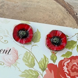 Peut inclure: Une paire de boucles d'oreilles en forme de coquelicot rouge avec des centres verts. Les boucles d'oreilles sont faites à la main et sont présentées sur une carte blanche à motif floral.