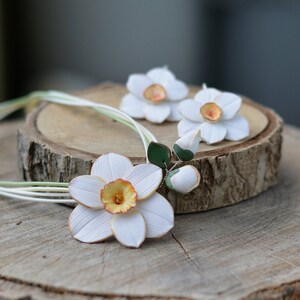 May include: A white paper flower necklace with three white paper daffodils, two of which are earrings. The necklace and earrings are on a wooden surface.