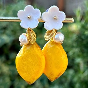 Könnte beinhalten: Ein Paar ohrringförmige Zitronen. Jeder Ohrring hat einen hellgelben Zitronenanhänger, eine weiße Blume, ein goldfarbenes Blatt und eine kleine Perle. Die Ohrringe werden auf einem goldfarbenen Ständer vor einem verschwommenen grünen Hintergrund präsentiert.