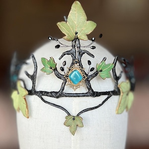 Puede incluir: Una corona artesanal con un diseño inspirado en la naturaleza. La corona presenta un marco oscuro en forma de rama adornado con hojas de color verde claro y una gema turquesa central. Las hojas están pintadas de color verde claro. La corona se exhibe en una cabeza de maniquí.