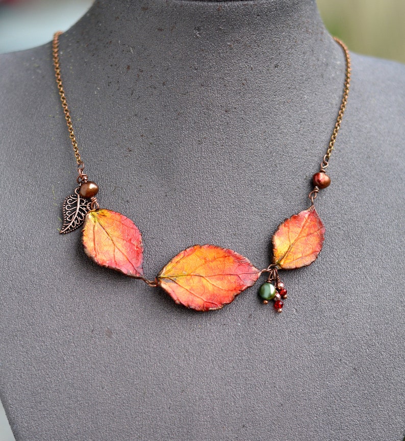 May include: A copper chain necklace with three orange, red, and yellow leaves, a small copper leaf charm, and a cluster of red and green beads.