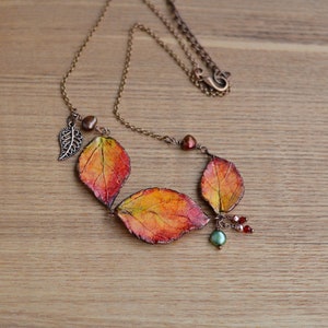 May include: A copper chain necklace with a pendant made of three orange, red, and yellow leaves. The leaves are attached to the chain with copper wire. There are also small copper beads and a single green bead on the chain.