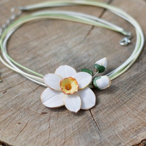 May include: A white and yellow polymer clay daffodil flower necklace with green leaves and buds. The necklace is made with multiple strands of white and green cord.