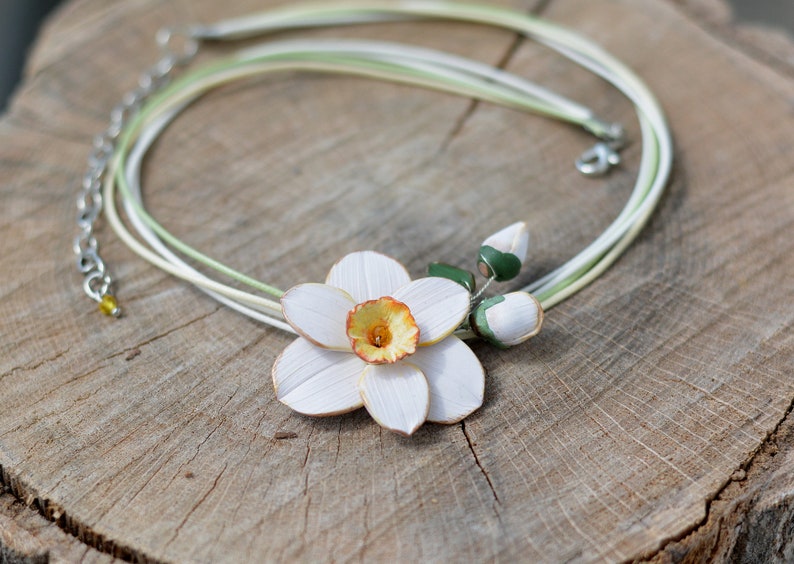 May include: A white and yellow polymer clay daffodil flower necklace with green leaves and buds. The necklace is made with multiple strands of white and green cord.