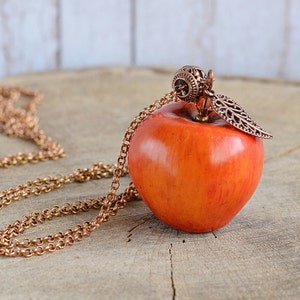 May include: A copper chain necklace with a red apple pendant. The apple has a detailed copper leaf and a decorative copper ball at the top.
