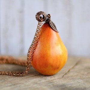 May include: A gold chain necklace with a pear-shaped pendant. The pendant is made of orange and yellow clay and has a small leaf charm attached to the top.