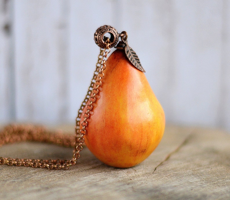 May include: A gold chain necklace with a pear-shaped pendant. The pendant is made of orange and yellow clay and has a small leaf charm attached to the top.