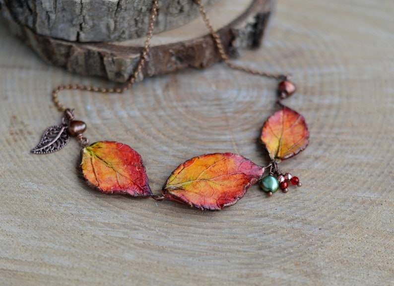 May include: A copper chain necklace with three orange, red, and yellow leaves, a small copper leaf charm, and a green and red bead cluster.