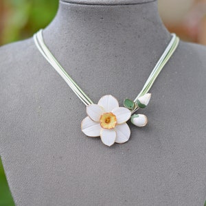May include: A white paper flower necklace with green leaves and a yellow centre. The necklace is made of multiple strands of white string.