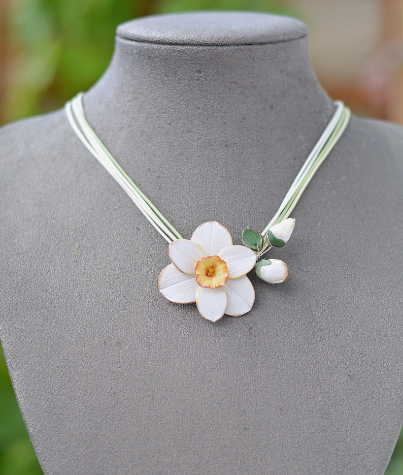 May include: A white paper flower necklace with green leaves and a yellow centre. The necklace is made of multiple strands of white string.