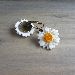 Op de afbeelding: Een paar madeliefjes oorbellen. Elke oorbel heeft een witte madeliefbloem met een geel centrum. De oorbellen hebben een bronzen metalen achterkant en een oorhaaksluiting. Op een lichtgrijze houten ondergrond.