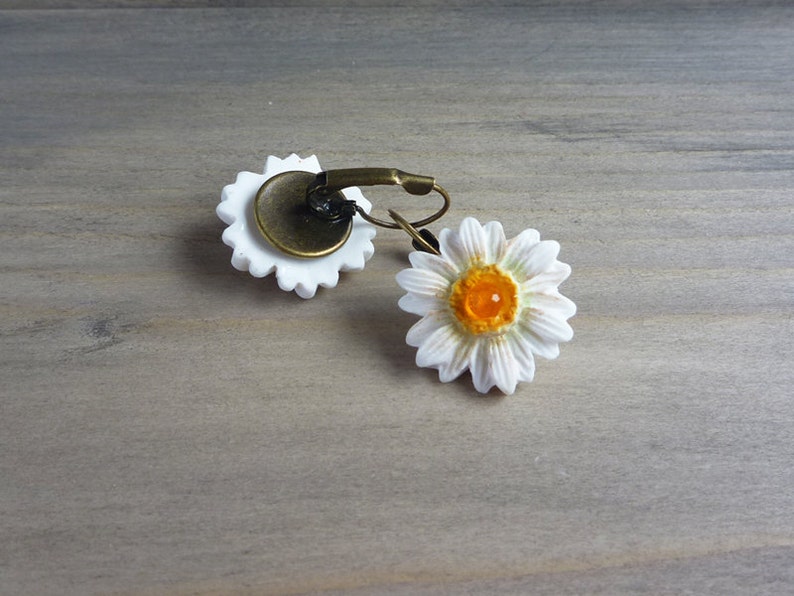 Op de afbeelding: Een paar madeliefjes oorbellen. Elke oorbel heeft een witte madeliefbloem met een geel centrum. De oorbellen hebben een bronzen metalen achterkant en een oorhaaksluiting. Op een lichtgrijze houten ondergrond.