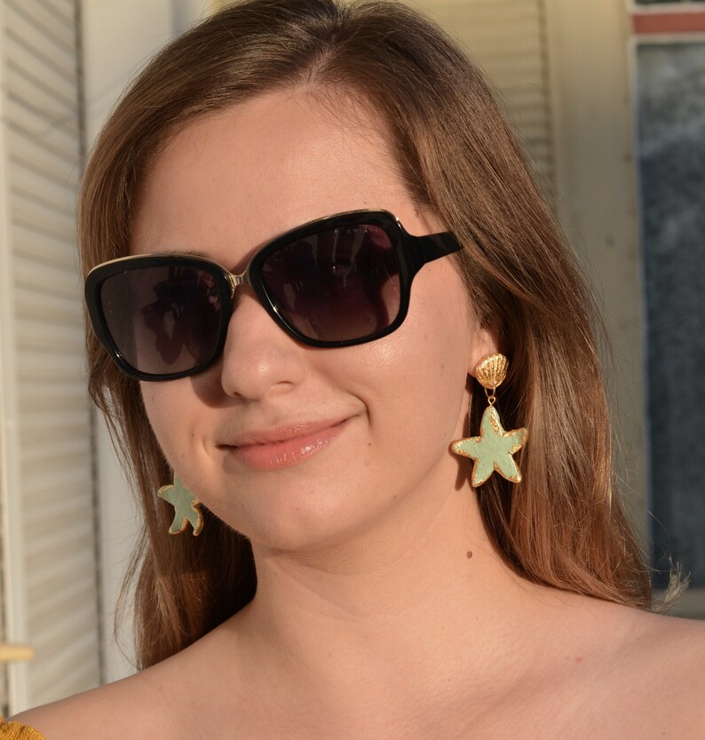 K&ouml;nnte beinhalten: Eine Frau tr&auml;gt eine schwarze Sonnenbrille mit goldenen Akzenten und ein Paar gr&uuml;ne und goldene Seestern-Ohrringe. Die Ohrringe haben einen kleinen Muschel-Anh&auml;nger &uuml;ber dem Seestern.