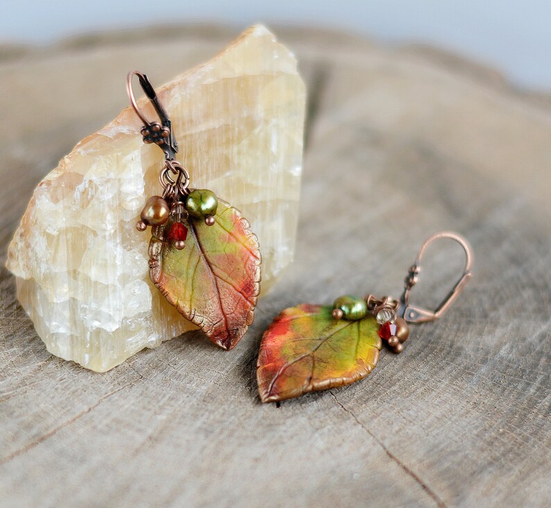 May include: A pair of earrings featuring copper leaf charms with red, green, and brown accents. The leaves are textured and have a realistic look.