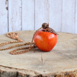 May include: A red apple pendant necklace with a copper chain and a decorative leaf charm. The necklace is displayed on a wooden surface.