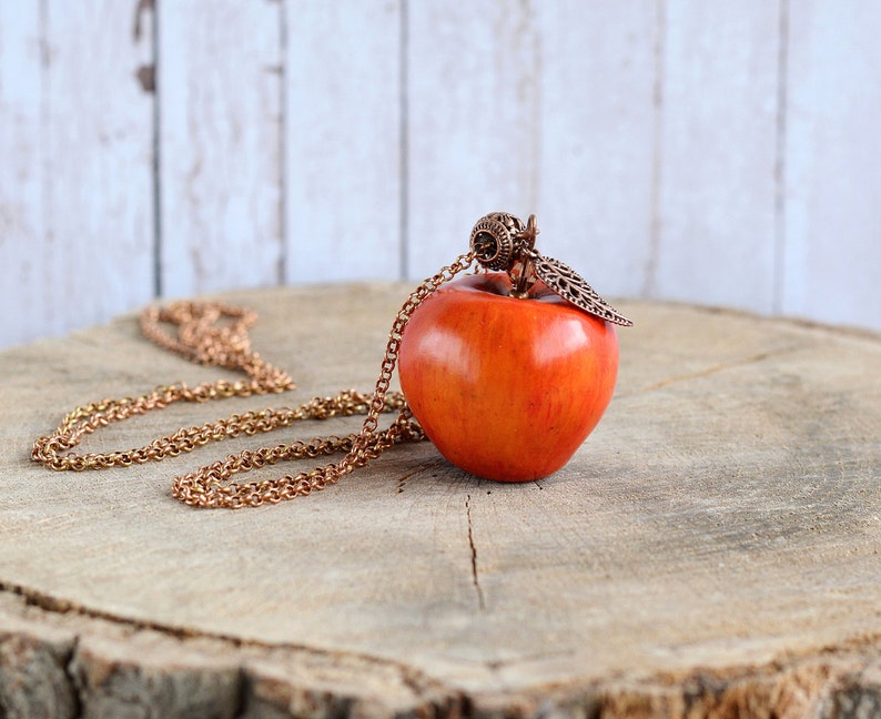 May include: A red apple pendant necklace with a copper chain and a decorative leaf charm. The necklace is displayed on a wooden surface.