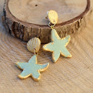 K&ouml;nnte beinhalten: Ein Paar goldene und hellblaue Seestern-Ohrringe mit einem goldenen Muschel-Anh&auml;nger. Die Ohrringe liegen auf einer Holzoberfl&auml;che.