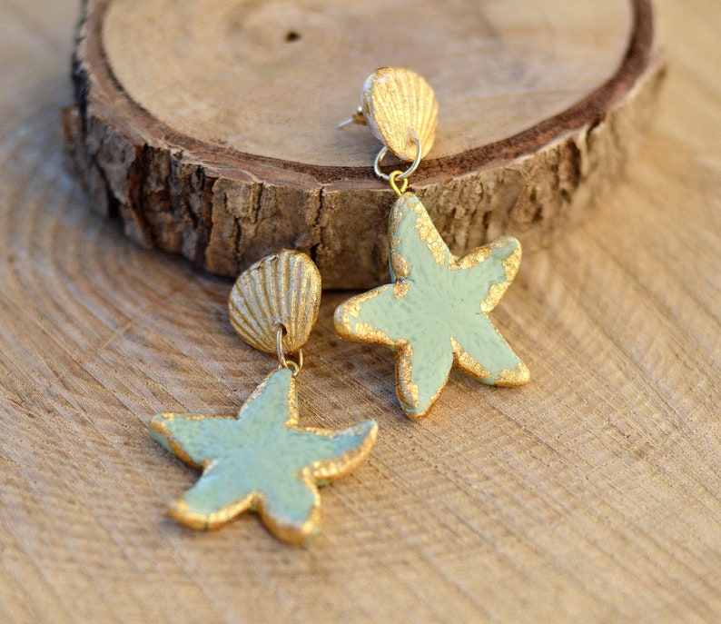 K&ouml;nnte beinhalten: Ein Paar goldene und hellblaue Seestern-Ohrringe mit einem goldenen Muschel-Anh&auml;nger. Die Ohrringe liegen auf einer Holzoberfl&auml;che.