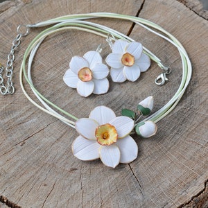 May include: A white and yellow daffodil flower necklace and earrings set. The necklace is made of multiple strands of white and green cord. The earrings are dangle earrings with a white daffodil flower.