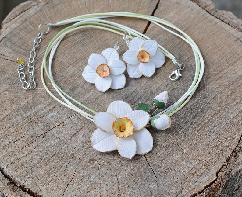 May include: A white and yellow daffodil flower necklace and earrings set. The necklace is made of multiple strands of white and green cord. The earrings are dangle earrings with a white daffodil flower.