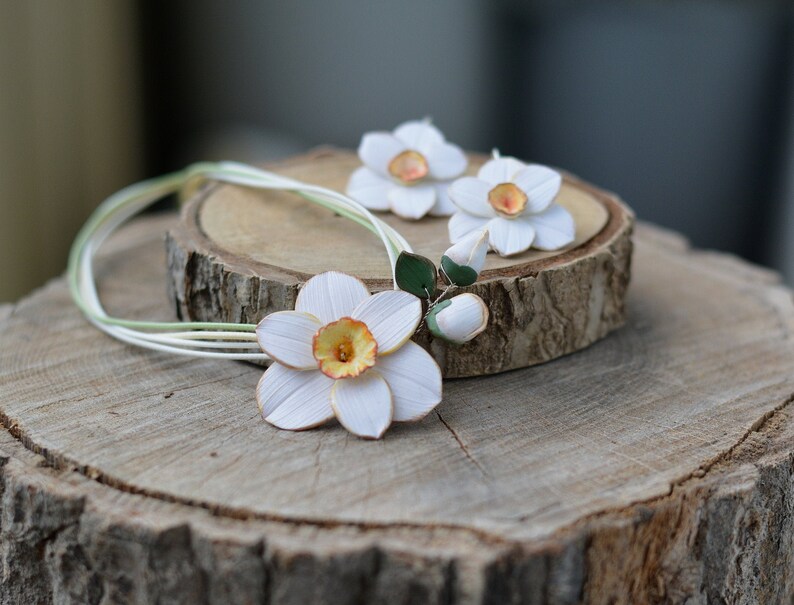 May include: A white paper flower necklace with three white paper daffodils, two of which are earrings. The necklace and earrings are on a wooden surface.