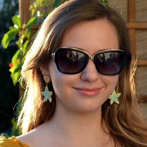 K&ouml;nnte beinhalten: Eine Frau tr&auml;gt eine schwarze Sonnenbrille mit goldenen Akzenten und ein Paar t&uuml;rkisfarbene und goldene Seestern-Ohrringe.