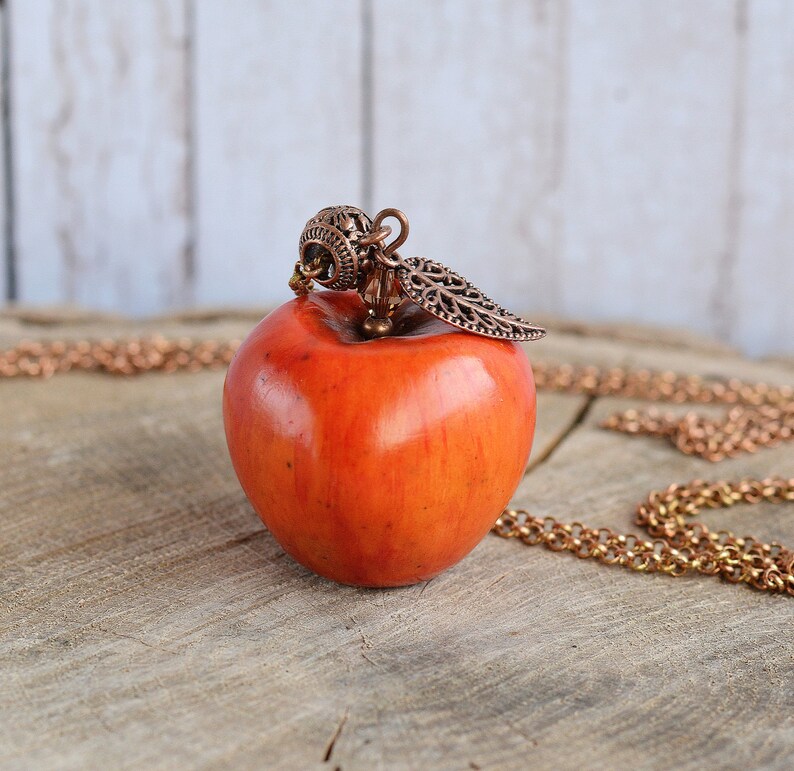 May include: A red apple pendant necklace with a copper leaf and a decorative copper bail. The necklace is on a wooden surface.