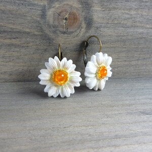 Op de afbeelding: Een paar madeliefjesoorbellen. Elke oorbel heeft een witte bloem met een getextureerd oppervlak en een feloranje centrum. De oorbellen hebben bronskleurige oorhaaksluitingen, tegen een rustieke houten achtergrond.