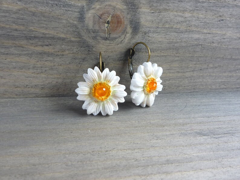 Op de afbeelding: Een paar madeliefjesoorbellen. Elke oorbel heeft een witte bloem met een getextureerd oppervlak en een feloranje centrum. De oorbellen hebben bronskleurige oorhaaksluitingen, tegen een rustieke houten achtergrond.