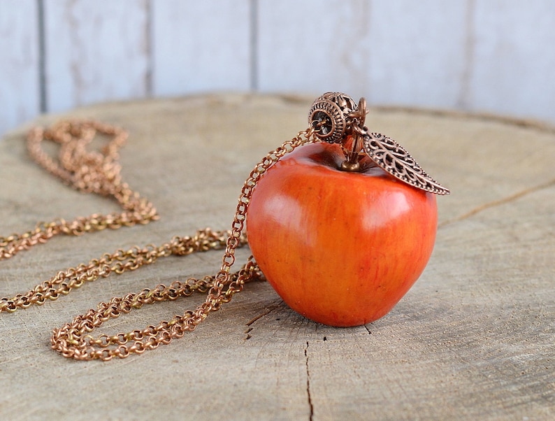 May include: A copper chain necklace with a red apple pendant. The apple has a detailed copper leaf and a decorative copper top.
