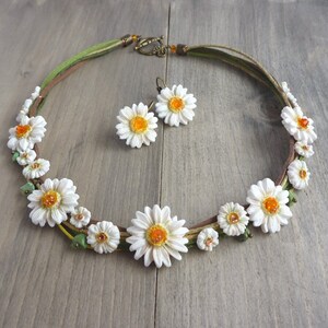 Op de afbeelding: Een handgemaakte ketting en oorbellen set met madelief bloem ontwerpen. De ketting heeft witte madeliefjes met oranje centra en groene details, geregen op een bruin en groen koord. De oorbellen passen bij het madelief ontwerp. De sieradenset wordt getoond op een houten oppervlak.