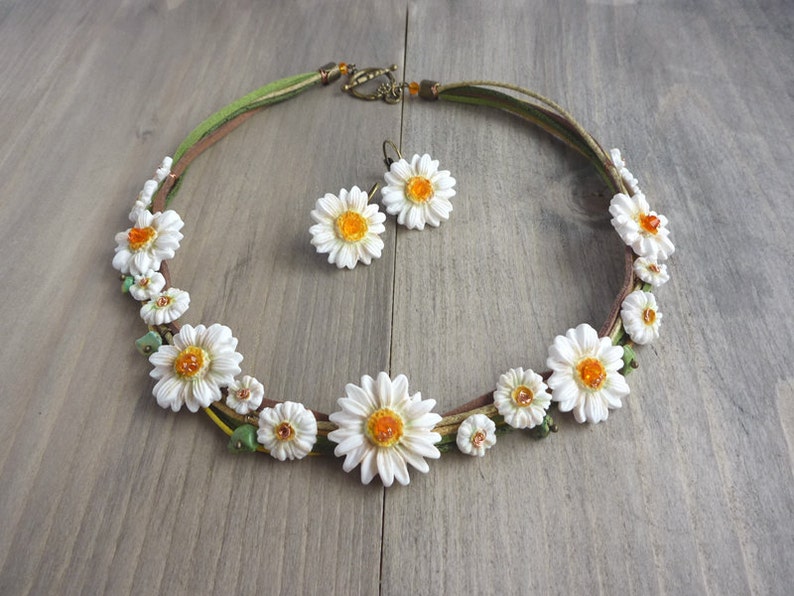 Op de afbeelding: Een handgemaakte ketting en oorbellen set met madelief bloem ontwerpen. De ketting heeft witte madeliefjes met oranje centra en groene details, geregen op een bruin en groen koord. De oorbellen passen bij het madelief ontwerp. De sieradenset wordt getoond op een houten oppervlak.