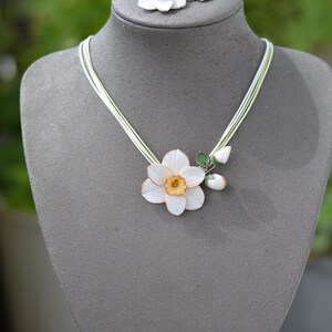 May include: A white porcelain necklace with a delicate daffodil flower pendant and matching earrings. The necklace features a single daffodil with a yellow centre and green leaves, while the earrings each have a single white daffodil.