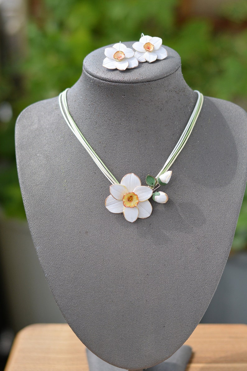 May include: A white porcelain necklace with a delicate daffodil flower pendant and matching earrings. The necklace features a single daffodil with a yellow centre and green leaves, while the earrings each have a single white daffodil.